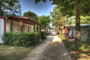 Campingplatz-Allee mit Zelten und Wohnwagen, Kiesweg zwischen Baeumen und Hecken, Motorrader und Fahrzeuge am Stellplatz.