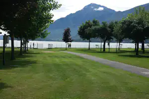 Campingplatz mit Rasenweg, Bäumen und Seeufer, im Hintergrund Berge und ein Zaun entlang des Wassers.