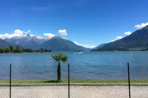 Blick auf den See mit Boot, Palmen vor einem Zaun und Bergpanorama im Hintergrund.