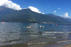 Seeufer mit Sandstrand, Schwimmern im Wasser und Bergen im Hintergrund, Boote auf dem See.