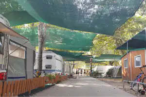 Wohnwagen und Zelte auf dem Campingplatz, zwischen Bäumen, mit grünen Sonnensegeln über dem Weg.