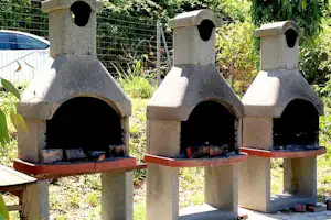 Drei gemauerte Grills mit offenen Feuerstellen auf einer Terrasse, umgeben von Hecken und Gartenpflanzen.