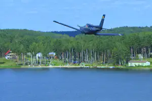 Flugzeug über einem Campingplatz am See, im Hintergrund Wald und mehrere Gebäude am Ufer.