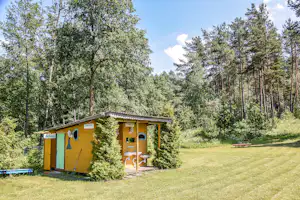 Gelbes Toilettenhäuschen mit grüner Tür und Schild „Tualetas“ auf dem Campingplatz, umgeben von Bäumen und Rasen.