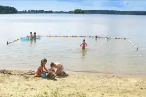 Badebereich am See: Kinder im Wasser vor bunten Schwimmringen, am Ufer sitzen zwei Personen im Sand.