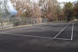 Asphalt-Tennisplatz auf dem Campingplatz mit Umzäunung, Linienmarkierungen und Bäumen im Hintergrund.