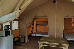 Innenansicht eines Glamping-Zelts mit Bett, Holztisch und Küchenzeile, seitlich offene Vorhänge und Zeltwände.