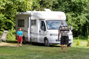 Wohnmobil am Campingplatz mit geöffneter Tür, daneben ein Kind und ein Erwachsener, im Hintergrund Bäume und Rasen.