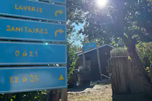Wegweiser am Campingplatz mit Schildern für Bar, Restaurant, Laverie und Sanitaires, dahinter Holzgebäude und Bäume.