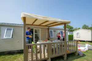 Mobilheim mit Veranda, Holzzaun und Tisch; zwei Personen bereiten am Grill vor, im Hintergrund weitere Wohnwagen.