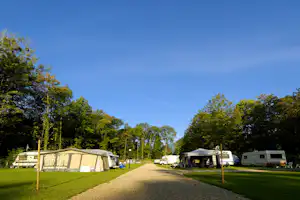 Zelte und Wohnwagen auf einem Campingplatz entlang eines Kieswegs im Wald, mit Bäumen und Rasenflächen.