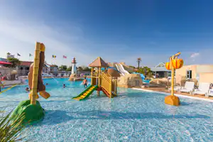 Familienpool mit Wasserrutsche, Spielgeräten und Liegestühlen am Beckenrand, im Hintergrund Gebäude und Palmen.
