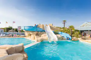 Blauer Wasserrutschenbereich am Pool mit Steinumrandung, Palmen und Liegen auf einem Campingplatz.