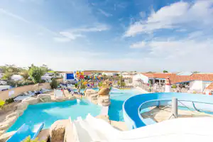 Blaue Camping-Pools mit Steinrand und Liegen, dahinter Terrassen und Gebäude unter klarem Himmel.