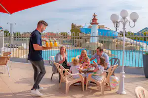 Familie am Tisch am Pool: Kinder und Erwachsene sitzen auf Stühlen, daneben ein Mann, im Hintergrund Zaun und Lampen.