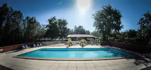Camping-Pool mit blauem Wasser, Stein-Terrasse, Liegestühlen und Zaun, im Hintergrund ein Gebäude und Bäume.