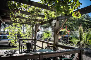Holzpergola mit Sitzgruppe aus Tisch und Stühlen, im Hintergrund Autos und Gartenpflanzen auf dem Campingplatz.