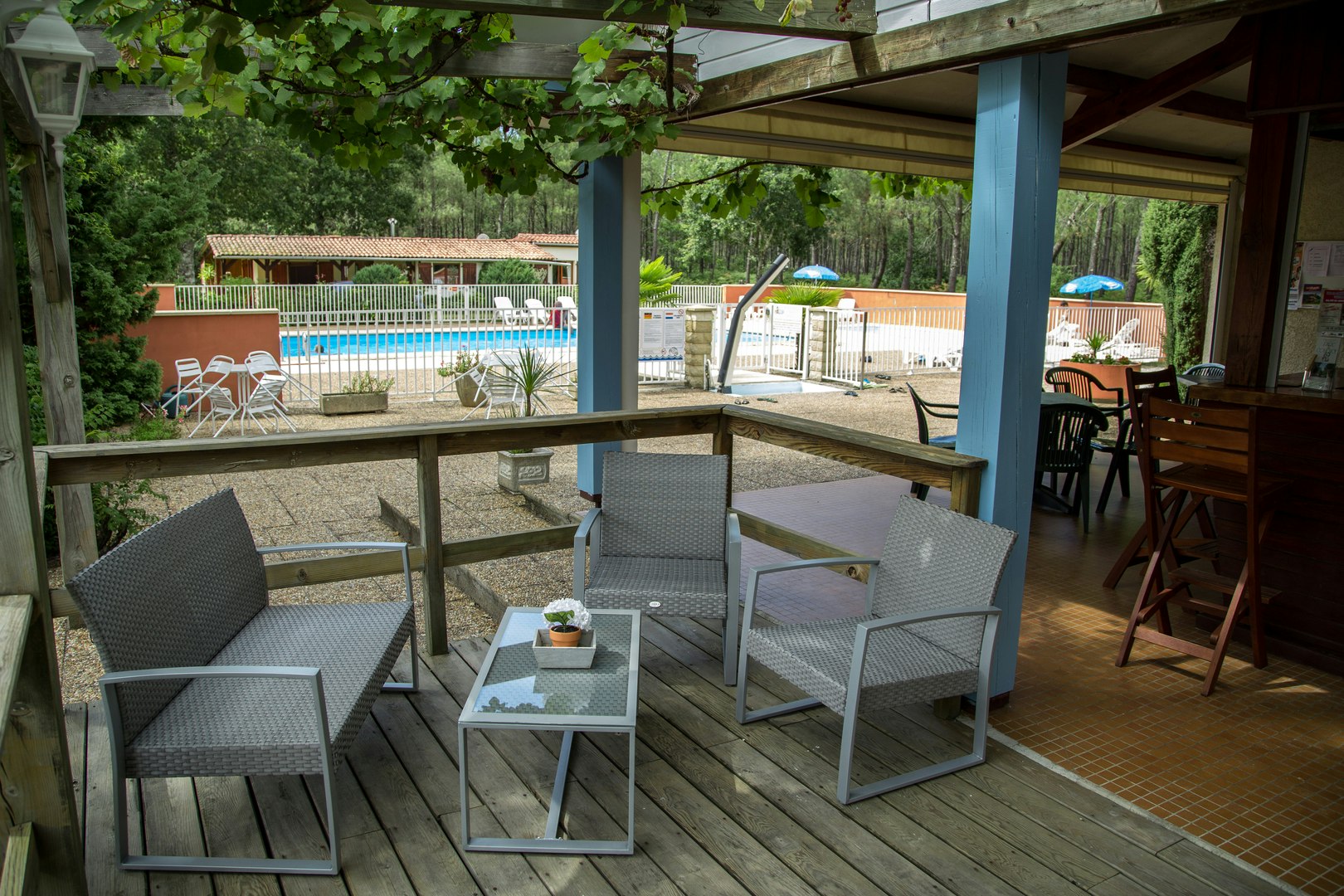 Sitzgruppe auf der Camping-Terrasse unter einem Dach, mit Tisch, Stühlen und Blick auf den Poolbereich und Bäume.