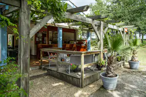 Terrasse am Campingplatz unter einer Holzpergola mit Sitzgruppe, Tisch und Pflanzen in Kübeln.