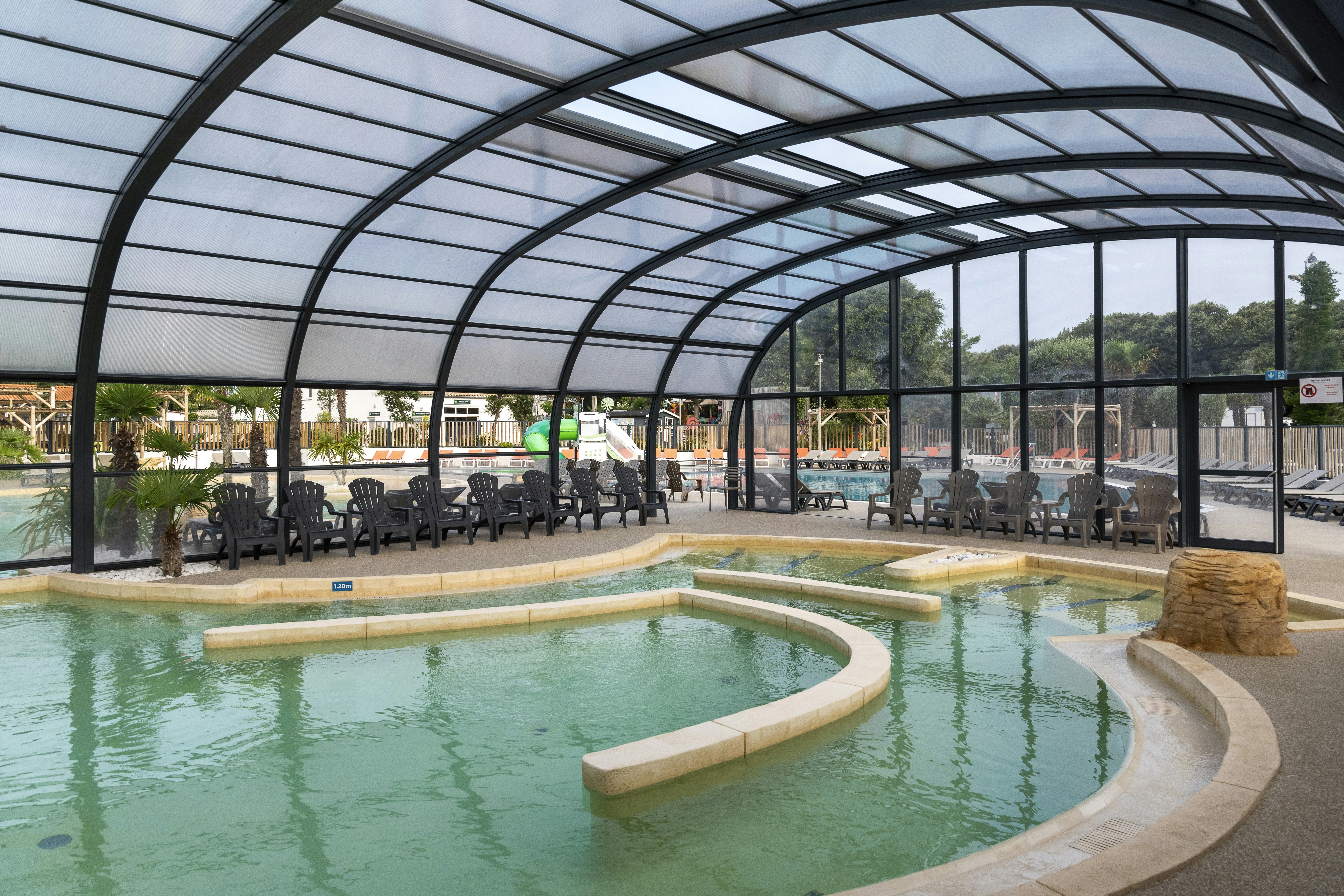 Innenpool mit rundem Becken unter Glasdach, Liegen und Tischen im Hintergrund, Blick auf Fenster und Außenbereich.