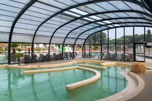 Innenpool mit rundem Becken unter Glasdach, Liegen und Tischen im Hintergrund, Blick auf Fenster und Außenbereich.