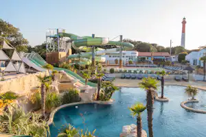 Großes Camping-Schwimmbecken mit Palmen, Rutschen und Liegen, im Hintergrund Gebäude und ein Leuchtturm.
