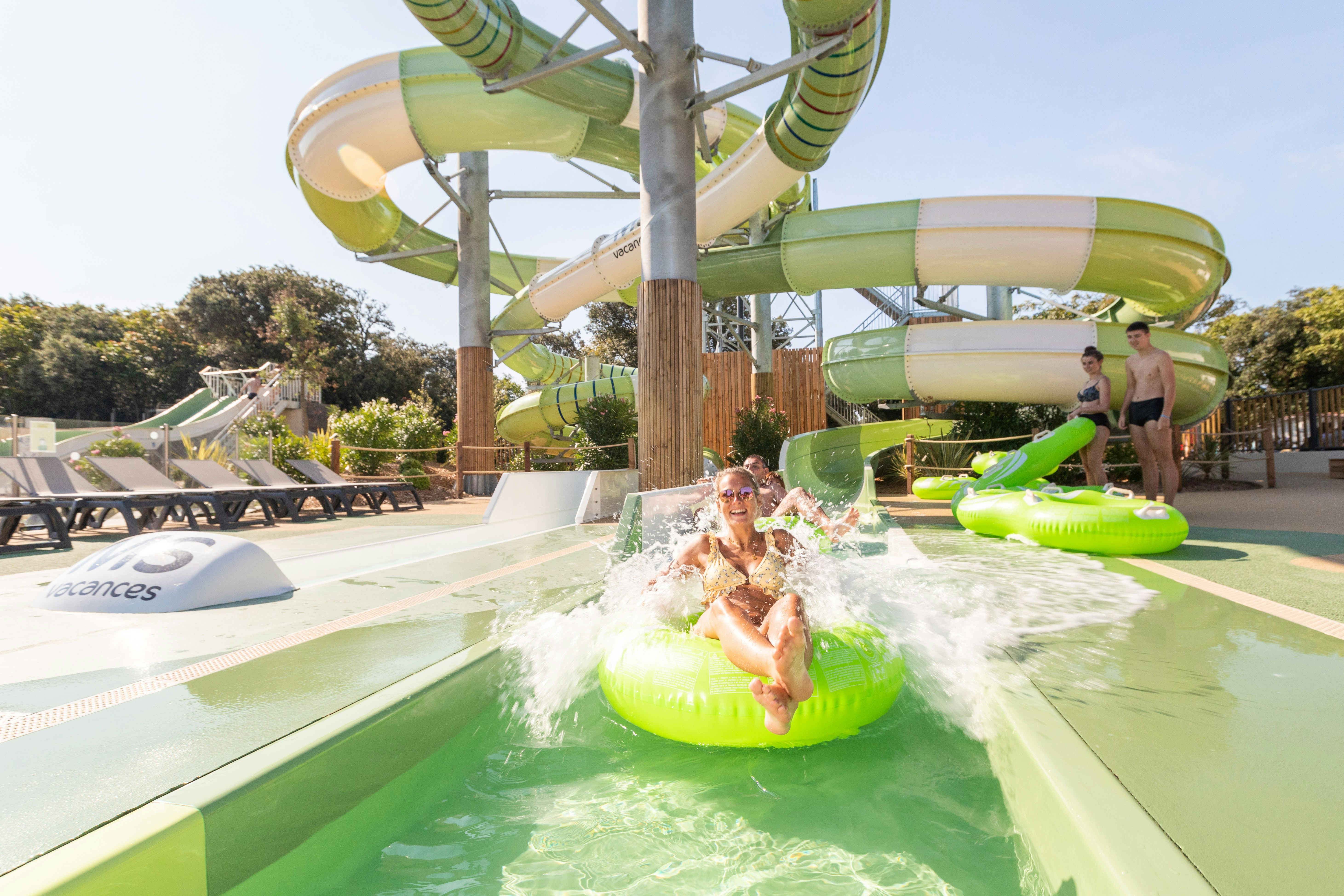 Grüner Wasserrutschen-Park mit Pool: Ein Kind rutscht ins Wasser, im Hintergrund Liegen und ein Betreuer am Beckenrand.