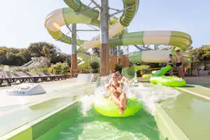 Grüner Wasserrutschen-Park mit Pool: Ein Kind rutscht ins Wasser, im Hintergrund Liegen und ein Betreuer am Beckenrand.