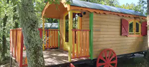 Holz-Wagenunterkunft mit Veranda, roter Tür und Fensterläden im Grünen, neben einem Baum und einem Kiesweg.