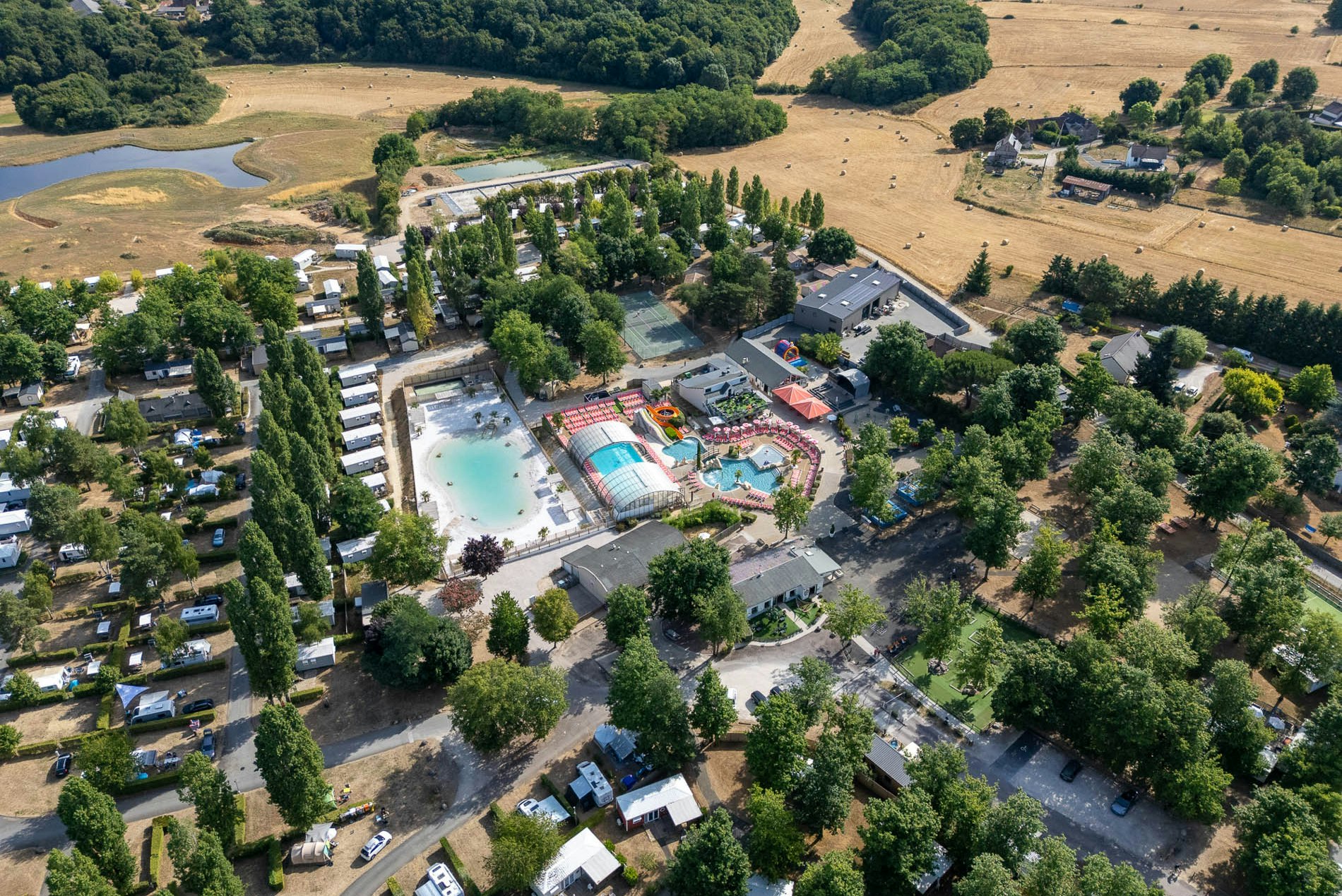 Luftaufnahme einer Campinganlage mit Schwimmbecken, beheiztem Hallenbereich, Sonnenterrassen und vielen Stellplätzen zwischen Bäumen.