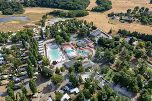 Luftaufnahme einer Campinganlage mit Schwimmbecken, beheiztem Hallenbereich, Sonnenterrassen und vielen Stellplätzen zwischen Bäumen.