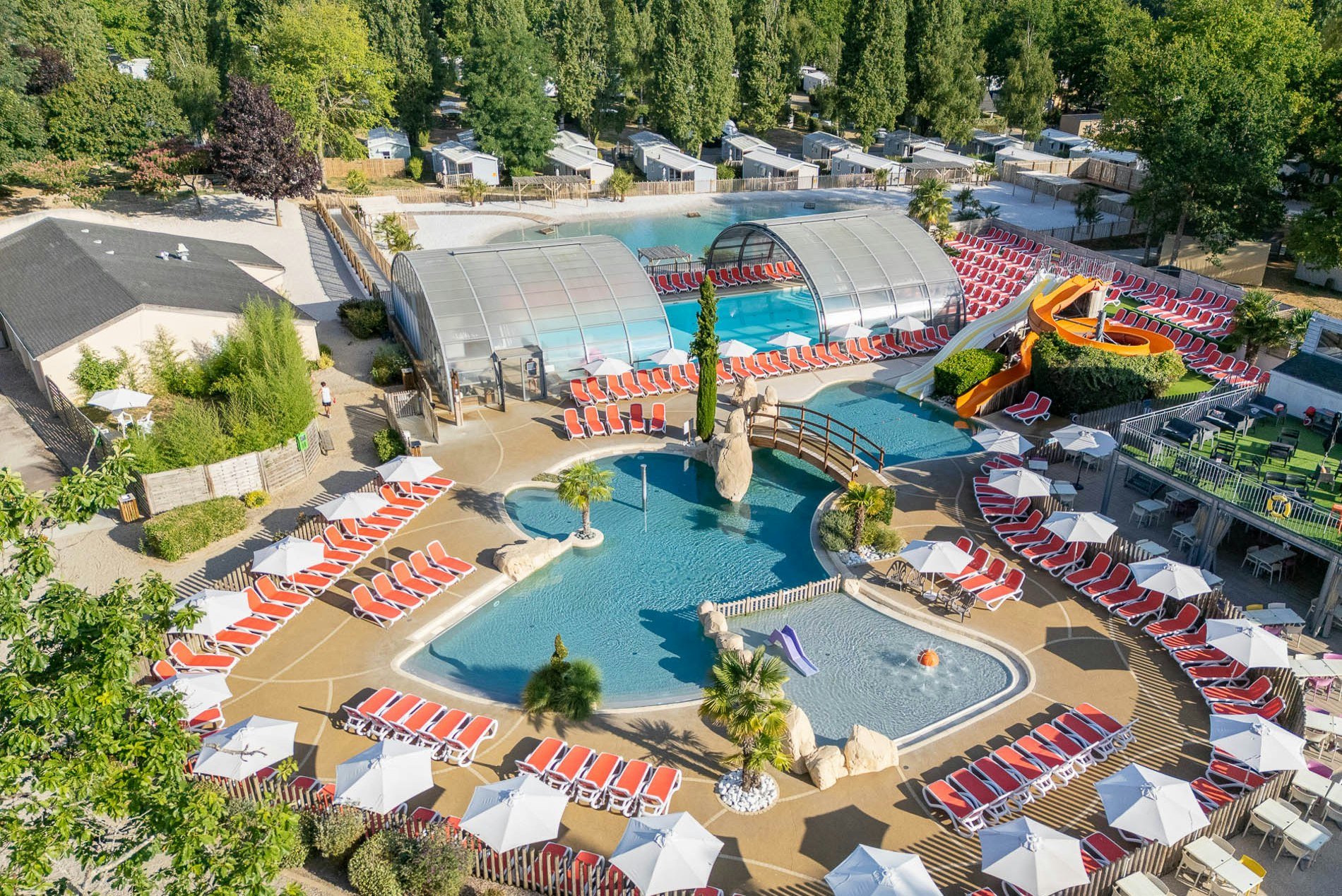 Schwimmbad im Campingbereich mit Sonnenschirmen, Liegestühlen, zwei Becken und einem großen Glasdach, umgeben von Bäumen.