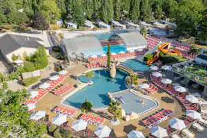 Schwimmbad im Campingbereich mit Sonnenschirmen, Liegestühlen, zwei Becken und einem großen Glasdach, umgeben von Bäumen.