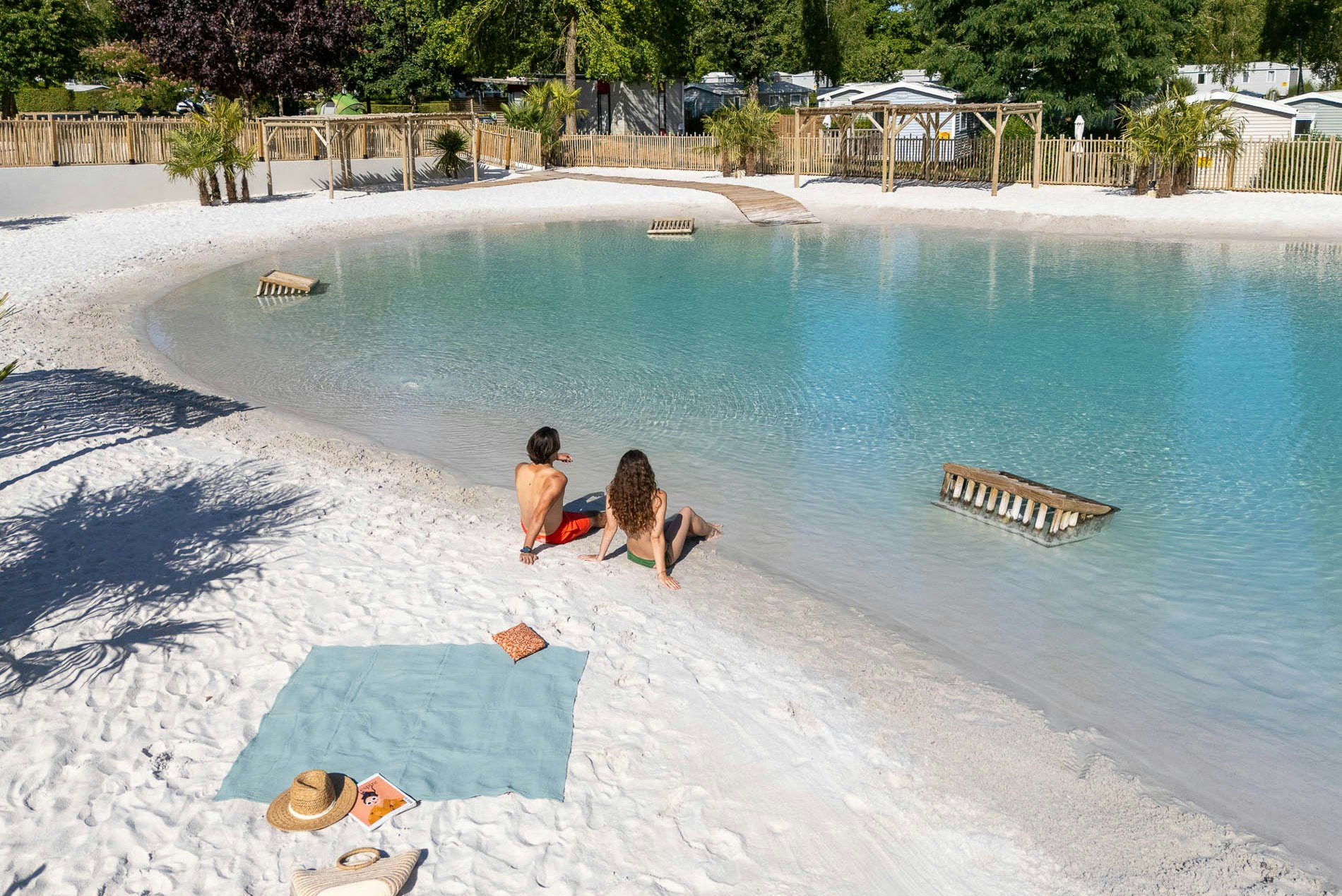 Campingpool mit türkisfarbenem Wasser, Holzliegen im Becken und Liegefläche am Rand, dahinter Zaun und Bäume.