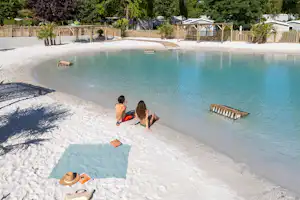 Campingpool mit türkisfarbenem Wasser, Holzliegen im Becken und Liegefläche am Rand, dahinter Zaun und Bäume.