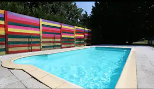 Blaues Camping-Poolbecken mit Rand aus Stein, daneben ein bunt gestreifter Zaun und Bäume im Hintergrund.