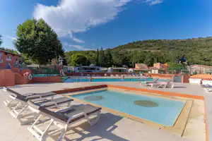 Camping-Pool mit eingelassenem Becken, Sonnenliegen und Terrasse, im Hintergrund Gebäude und Bäume vor einem bewaldeten Hügel.