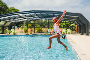Kind springt im Schwimmbecken eines Campingplatzes, überdachtes Pooldach und Liegen im Hintergrund.