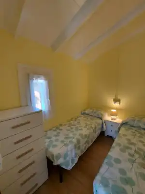 Schlafzimmer mit zwei Einzelbetten, gemusterten Decken, Nachttisch und Lampe, gelben Wänden und offener Tür zum Fensterbereich.