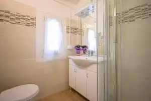 Modernes Badezimmer mit Waschbecken, Schrank, Spiegel und Dusche, hell gefliest, mit Fenster und Handtuchhalter.