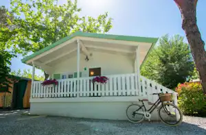 Ferienhaus mit Veranda, weißem Geländer und Fahrrad am Eingang, umgeben von Bäumen und gepflegtem Gartenweg.