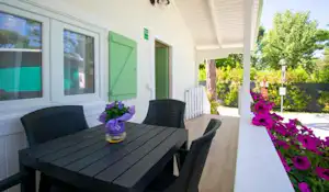 Camping-Terrasse mit schwarzem Tisch und Stühlen, grüner Tür, Blumenbeeten und Sicht auf Bäume neben dem Wohnbereich.