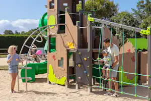 Kletterturm mit Rutsche und Seilnetz, Kinder spielen auf dem Spielplatz am Campingplatz, im Hintergrund Bäume und Rasen.