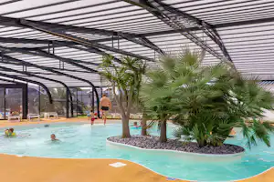Überdachter Innenpool mit Palmen im Beckenbereich, Sonnenschirme und Liegen unter einem Glasdach.