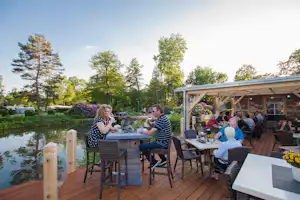 Terrasse am Campingplatz am See mit Holztischen, Stühlen und einem Restaurant unter Holzüberdachung, im Hintergrund Bäume und Blumen.