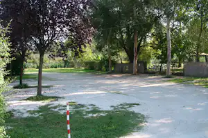 Campingplatz mit Kiesweg und Rasenfläche, umgeben von Bäumen und Hecken, im Hintergrund Holzzäune und ein Gebäude.