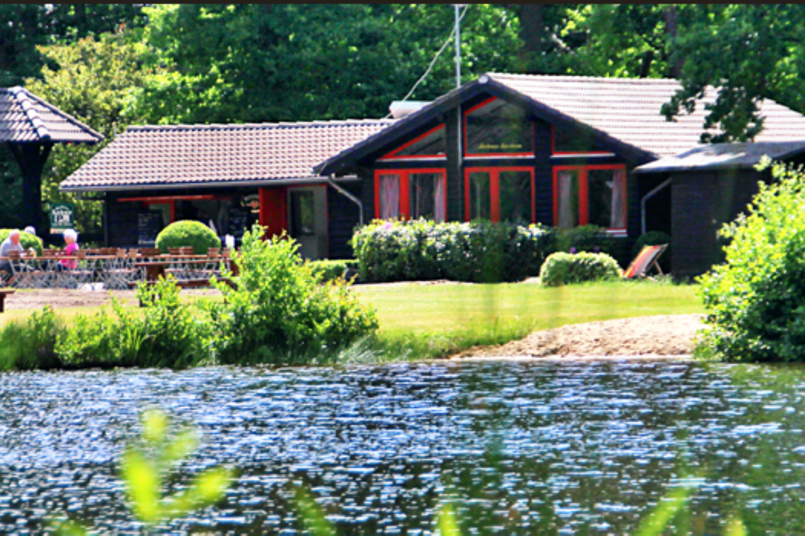 Campingplatz mit einem roten Holzbungalow und gepflegtem Garten, umgeben von Hecken und einem kleinen Teich mit Seerosen.