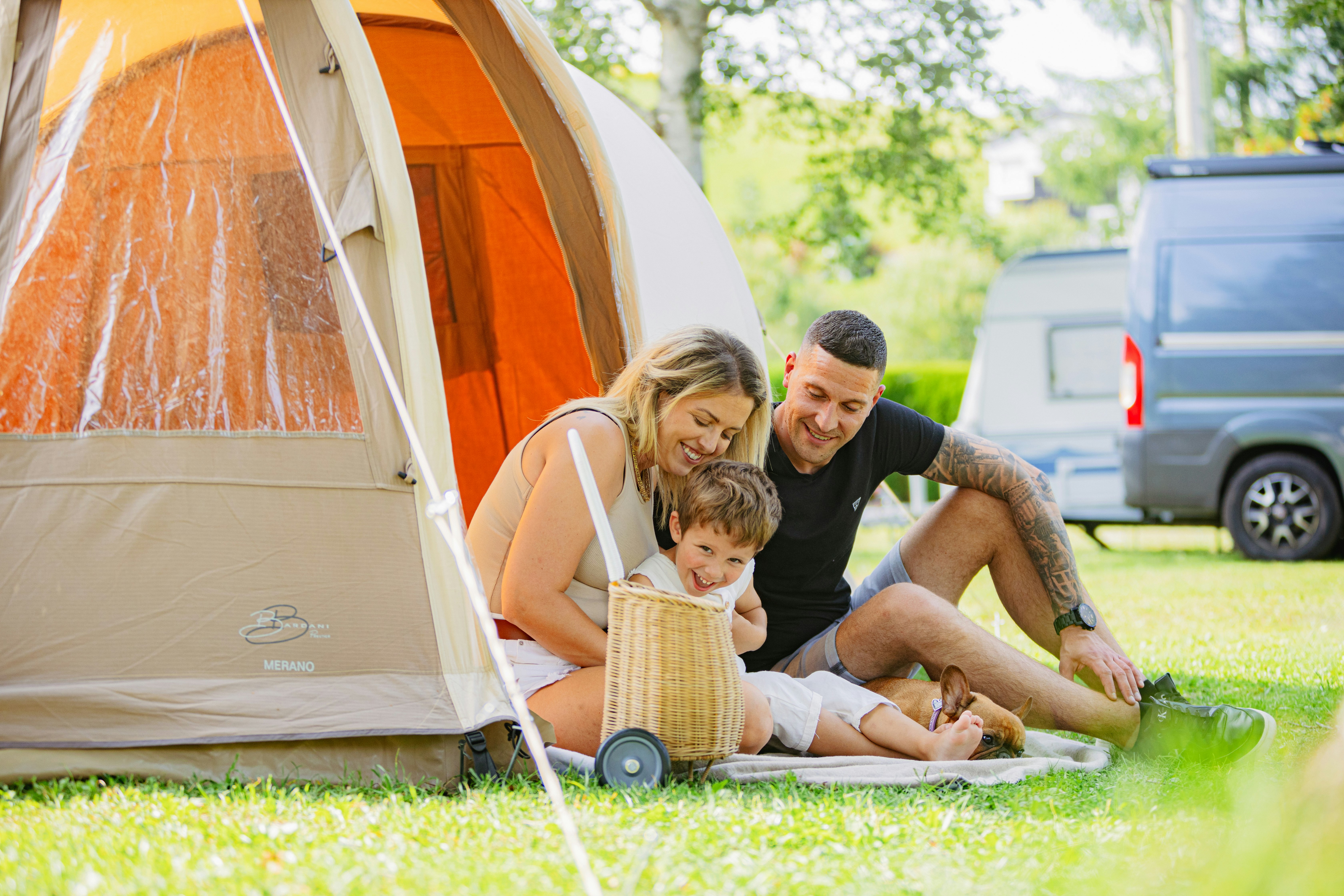 Familie sitzt vor einem Zelt auf dem Campingplatz, Kind spielt mit Spielzeug auf der Wiese, daneben steht ein Auto.