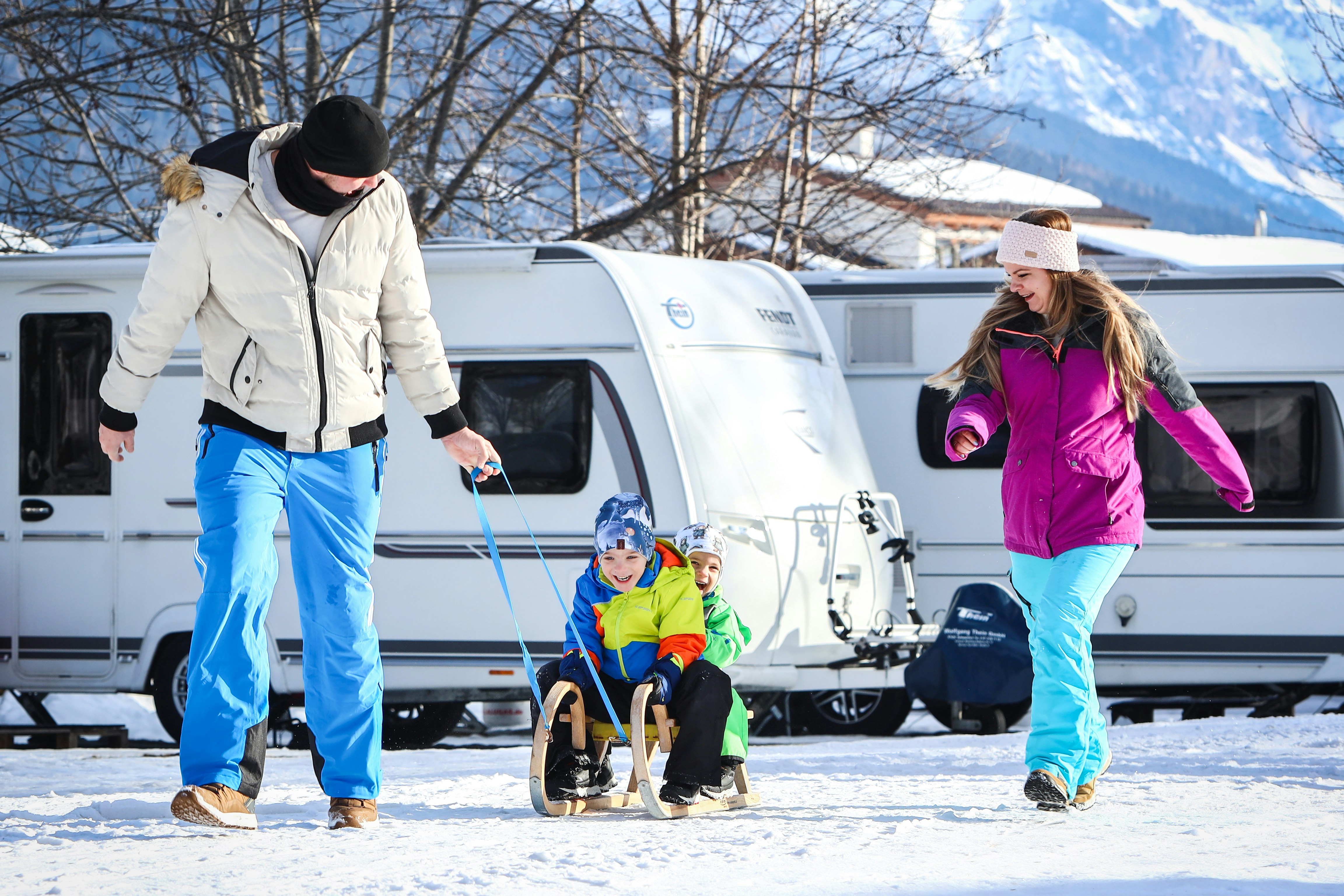 Zwei Personen stehen vor einem Wohnmobil auf verschneitem Campingplatz, im Hintergrund Bäume und Berge.