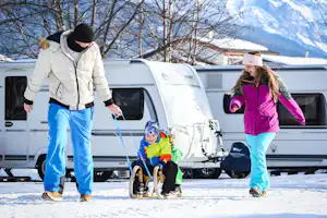 Zwei Personen stehen vor einem Wohnmobil auf verschneitem Campingplatz, im Hintergrund Bäume und Berge.